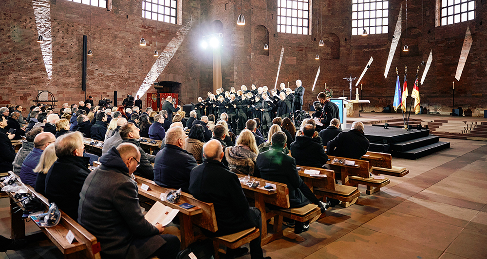Zahlreiche Menschen sitzen auf Holzbänken in einer großen Halle. Vor ihnen steht ein singender Chor auf einer erhöhten Bühne.