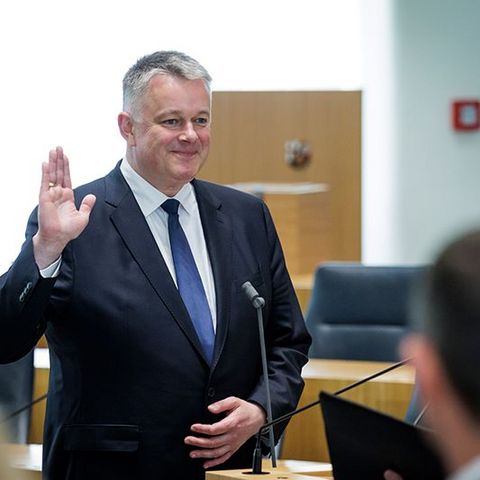 Herr Schnieder steht mit erhobener rechter Hand vor einem Mikrofon.
