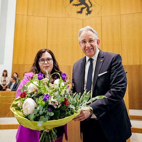 Jaqueline Rauschkolb und Matthias Lammert stehen nebeneinander im Plenarsaal und halten gemeinsam einen großen Blumenstrauß. Im Hintergrund sitzen mehrere Personen.