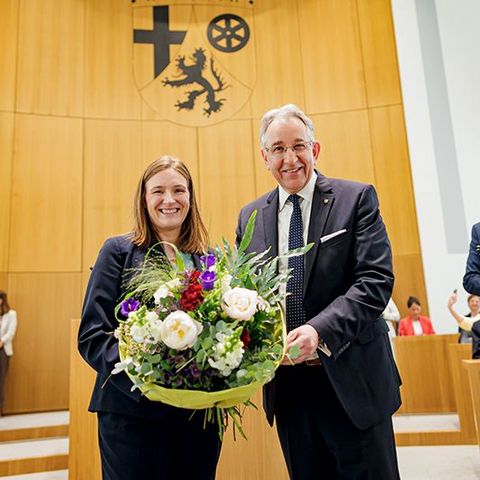 Katharina Binz und Matthias Lammert stehen nebeneinander im Plenarsaal und halten gemeinsam einen großen Blumenstrauß. Im Hintergrund sitzen mehrere Personen.