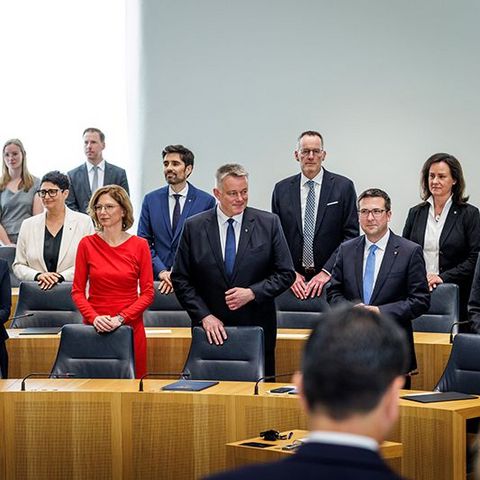 Die Staatsministerinnen und Staatsminister stehen gemeinsam mit Gordon Schnieder hinter ihren Plätzen im Plenarsaal.