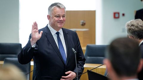 Herr Schnieder steht mit erhobener rechter Hand vor einem Mikrofon.