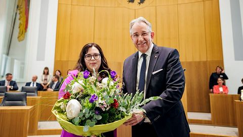 Jaqueline Rauschkolb und Matthias Lammert stehen nebeneinander im Plenarsaal und halten gemeinsam einen großen Blumenstrauß. Im Hintergrund sitzen mehrere Personen.