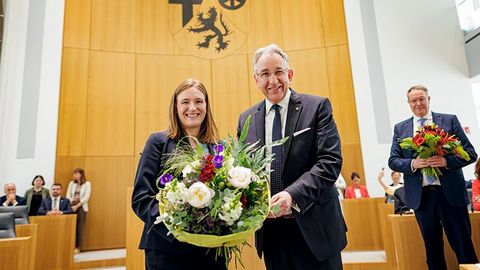 Katharina Binz und Matthias Lammert stehen nebeneinander im Plenarsaal und halten gemeinsam einen großen Blumenstrauß. Im Hintergrund sitzen mehrere Personen.