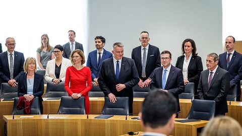 Die Staatsministerinnen und Staatsminister stehen gemeinsam mit Gordon Schnieder hinter ihren Plätzen im Plenarsaal.