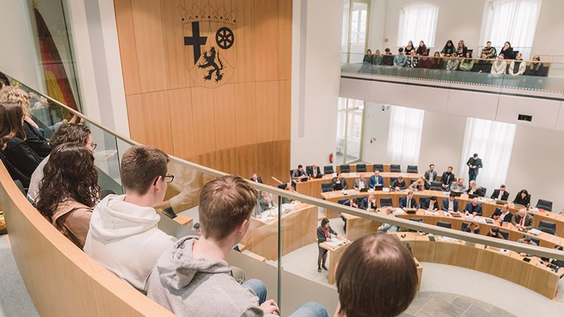 Zu sehen sind auf der linken Seite einige Personen, die auf der Besuchertribüne sitzen. Auf der rechten Seite des Bildes sieht man die Abgeordneten des Landtags, die auf ihren Plätzen im Plenarsaal sitzen.