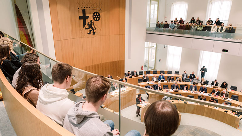 Die Besuchertribüne im Plenarsaal des rheinland-pfälzischen Landtags: eine Jugengruppe hört konzenriert der Debatte der Abgeordneten zu; der Blick von oben auf das Plenum und im Hintergrund die Holvertäfelung mit dem rheinland-pfälzischen Wappen