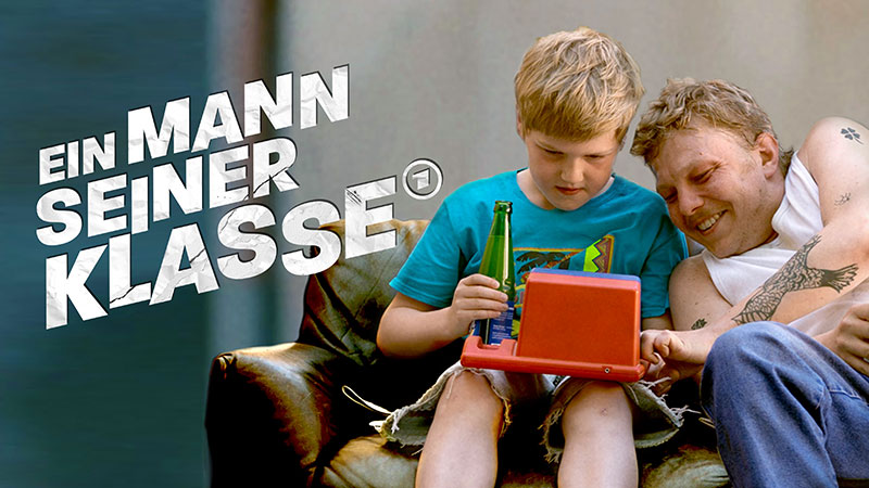 Vater und Sohn sitzen gemeinsam freudestrahlend auf einer Couch. Der hat ein Spielzeug auf dem Schoß. Das Bild trägt den Titel des Films: "Ein Mann seiner Klasse"