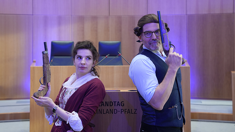 Zwei Personen in historischer Kleidung stehen vor dem Redepult des Landtags und halten Pistolenattrappen in den Händen.
