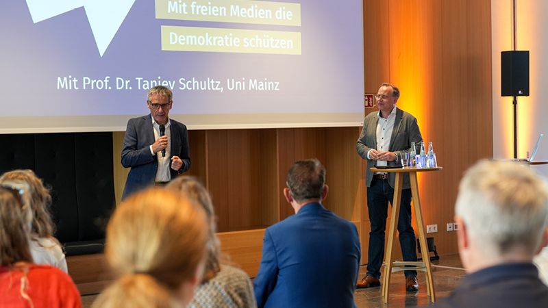 Zu sehen sind zwei Männer. Sie stehen im Restaurant des Landtags vor einer großen Leinwand. Auf der Leinwand steht: "#4 Tu was! Mit freien Medien die Demokratie schützen, mit Prof. Dr. Tanjev Schultz". Im Vordergrund sitzt das Publikum. Der Mann auf der rechten Seite steht hinter einem Stehtisch. Auf diesem stehen Wasserflaschen. Der Mann auf der linken Seite hält ein Mikrofon in der Hand und spricht zum Publikum.