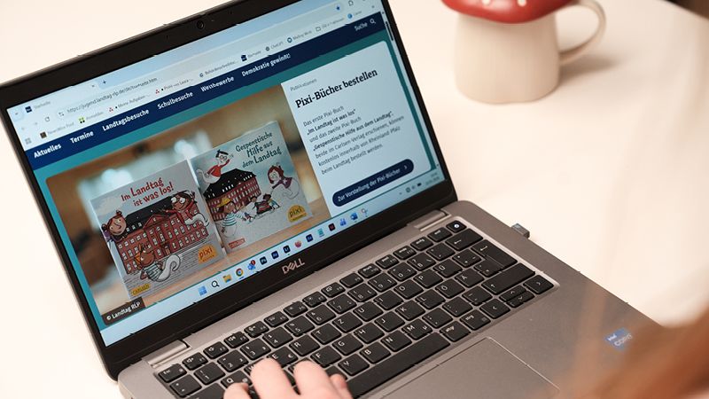 Zu sehen ist ein Laptop, der auf dem Tisch steht. Eine Person benutzt diesen gerade und tippt auf der Tastatur. Auf dem Bildschirm ist eine Website geöffnet. Man erkennt zwei Pixi-Bücher. Neben dem Laptop steht eine Tasse mit einem roten Deckel, der wie ein Pilz gestaltet ist.