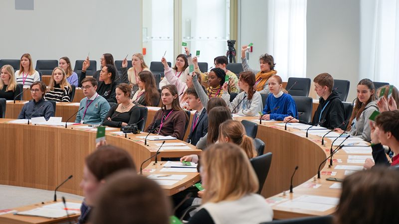 Zu sehen sind mehrere Schüler. Sie sitzen im Plenarsaal und hören gespannt zu.