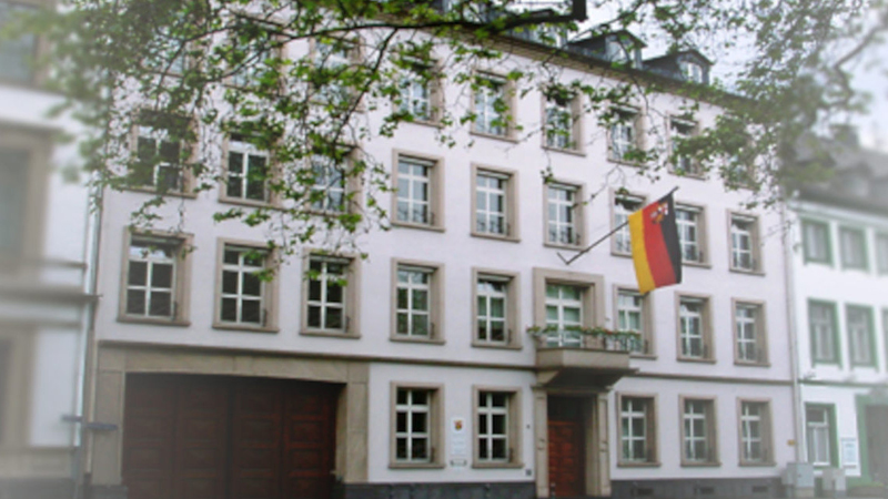 Zu sehen ist ein mehrst&ouml;ckiges, helles Geb&auml;ude mit vielen Fenstern. An der Fassade h&auml;ngt eine deutsche Flagge, die von einem kleinen Balkon &uuml;ber dem Eingangsbereich ausgeht.