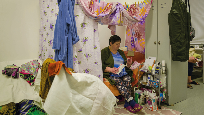 Zu sehen ist eine Frau. Sie sitzt auf ihrem Bett. Dieses steht in einem kleinen Raum. Über dem Bett wurden viele bunte Decken mit Wäscheklammern befestigt. Rechts neben dem Bett steht ein schmaler grauer Schrank.