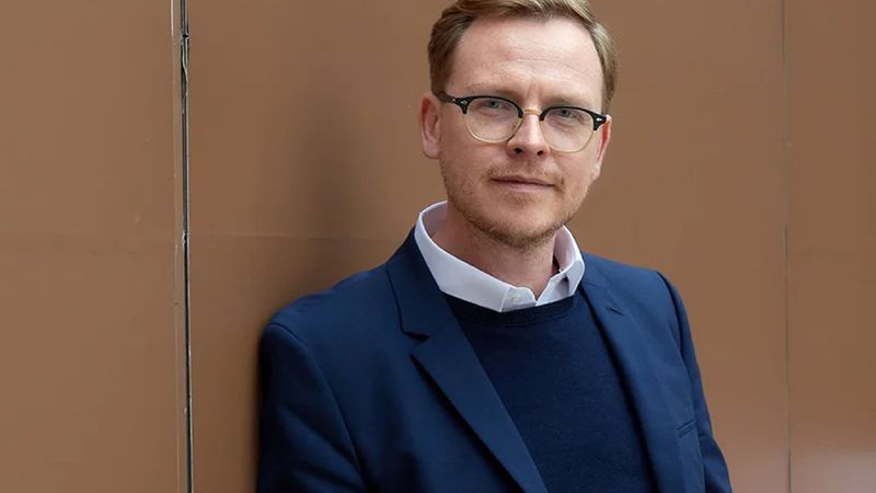 Zu sehen ist ein Mann mit einer Brille. Der Mann tr&auml;gt einen dunkelblauen Anzug. Er schaut in die Kamera. Der Mann steht vor einer hellbraunen Wand.