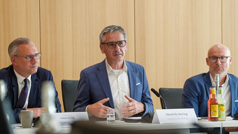 Zu sehen sind drei Männer. Sie sitzen im Saal des Landtags Rheinland-Pfalz. Alle drei Personen tragen einen blauen Anzug. Sie unterhalten sich miteinander.