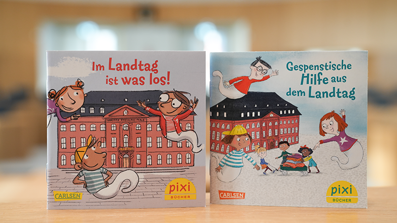 Zu sehen sind die Cover der zwei Pixi-Bücher aus dem Landtag. Darauf zu erkennen sind gezeichnete Figuren. Im Hintergrund sieht man den Plenarsaal leicht verschwommen.