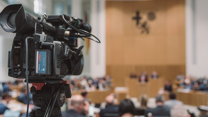 Zu sehen ist eine Kamera auf der linken Seite des Bildes. Im Hintergrund sieht man den Plenarsaal.