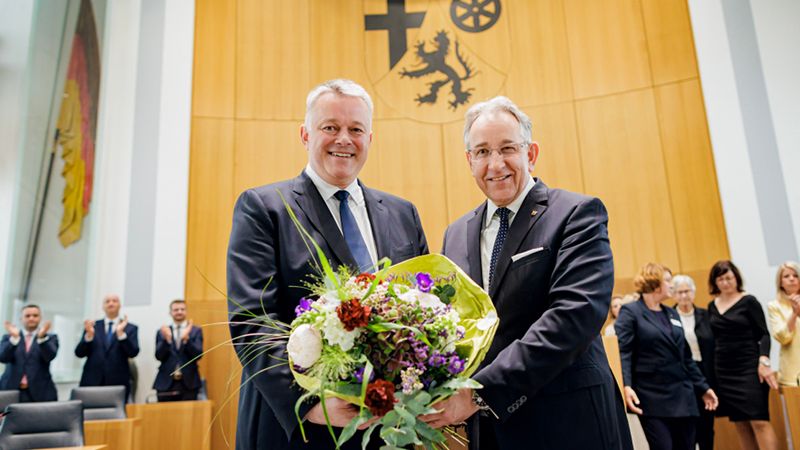 Das Bild zeigt zwei Männer in Anzügen im Plenarsaal. Gemeinsam halten sie einen großen Blumenstrauß und lächeln in die Kamera.
Im Hintergrund sieht man das Wappen von Rheinland-Pfalz an der Wand, eine deutsche Flagge links und mehrere applaudierende Personen.