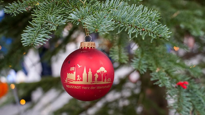 Zu sehen ist eine rote Weihnachtsbaumkugel. Aud dieser Kugel steht "Neustadt Herz der Weinstraße". Sie hängt an einem grünen Tannenbaum. Im Hintergrund sieht man Lichter.