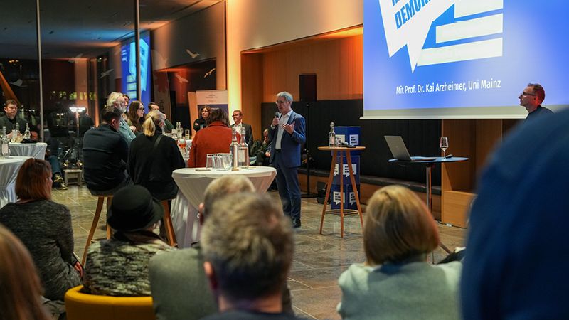 Zu sehen sind viele Personen. Sie sitzen an Tischen im Esszimmer des Landtags und hören gespannt dem Redner zu. Dieser steht vor einer großen blauen Leinwand mit dem Schriftzug "Werte. Haltung. Demokratie".