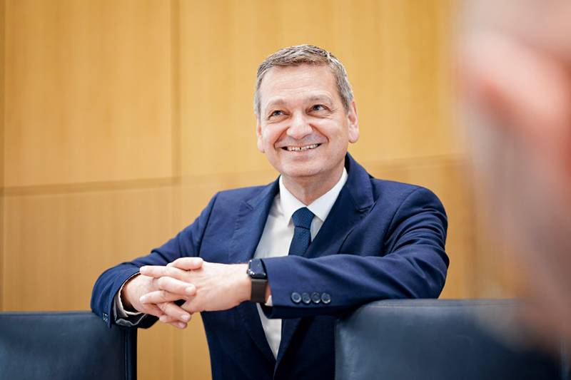 Zu sehen ist ein Mann. Er trägt einen dunkelblauen Anzug und steht hinter zwei Stühlen im Plenarsaal. Er lächelt.