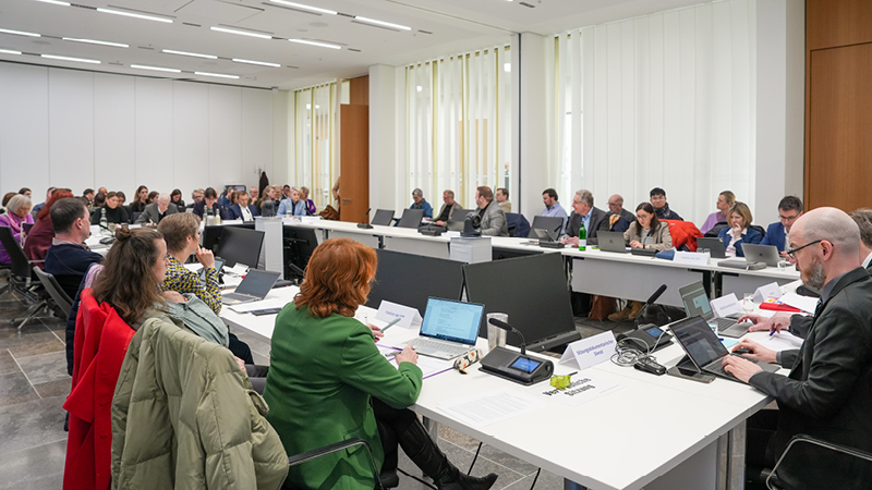 Zu sehen ist ein gro&szlig;er Konferenzraum, in dem viele Menschen an U-f&ouml;rmig angeordneten Tischen sitzen. Die Teilnehmenden haben Laptops, Unterlagen und Mikrofone vor sich.