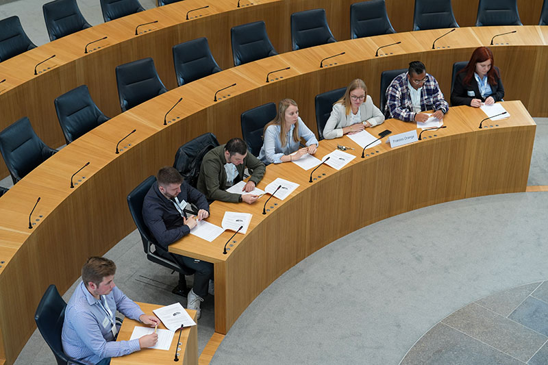 Die Teilnehmenden sitzen während den Debatten auf den Stühlen der Abgeordneten im Plenarsaal (Vogelperspektive).