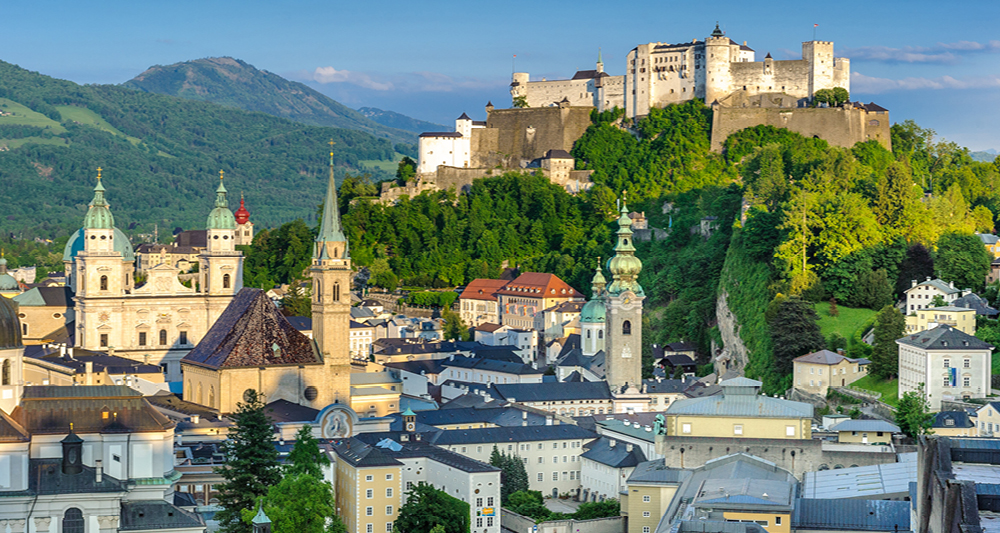 Auf dem Bild sieht man die Altstadt von Salzburg. Im Vordergrund stehen barocke Gebäude und mehrere Kirchen, darunter der beeindruckende Salzburger Dom mit seinen markanten Kuppeln.
Auf dem Hügel im Hintergrund thront die große Festung "Festung Hohensalzburg"
