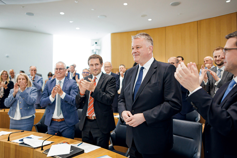 Viele Personen stehen im Plenarsaal. In der Mitte steht ein Mann im dunklen Anzug, während die Menschen um ihn herum applaudieren. 