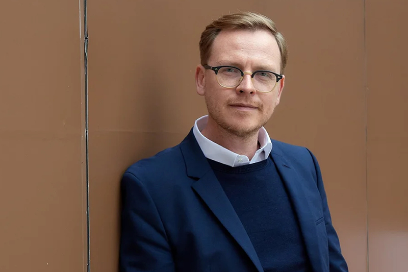 Zu sehen ist ein Mann mit einer Brille. Der Mann trägt einen dunkelblauen Anzug. Er schaut in die Kamera. Der Mann steht vor einer hellbraunen Wand.