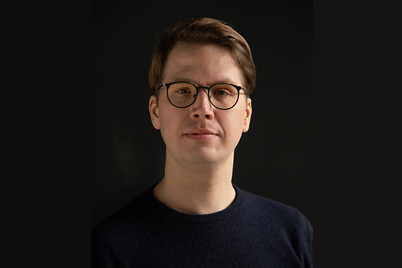 Zu sehen ist ein Mann. Er trägt eine schwarze Brille und schaut direkt in die Kamera. Der Hintergrund ist dunkel.