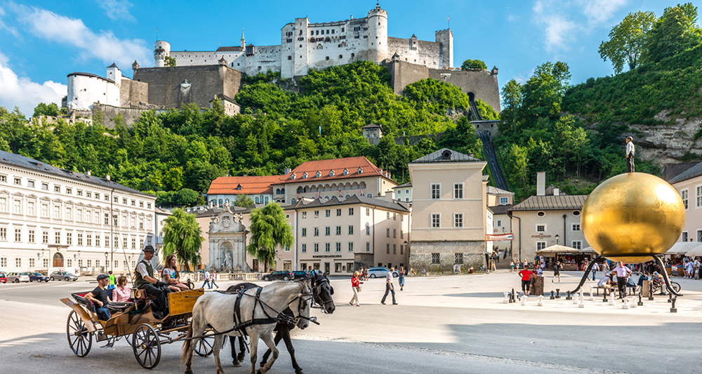 Zu sehen ist ein bekannter Platz in der Altstadt von Salzburg. Im Hintergrund thront die große Festung Hohensalzburg auf einem bewaldeten Hügel. Im Vordergrund befindet sich ein offener Platz mit historischen Gebäuden. Rechts steht die goldene Kugel-Skulptur Sphaera. Links sieht man eine Pferdekutsche mit Fahrgästen.