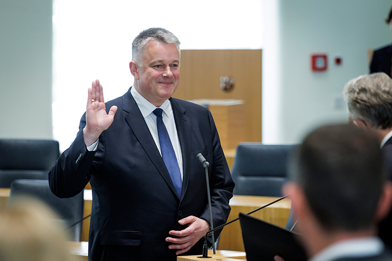 Herr Schnieder steht mit erhobener rechter Hand vor einem Mikrofon.