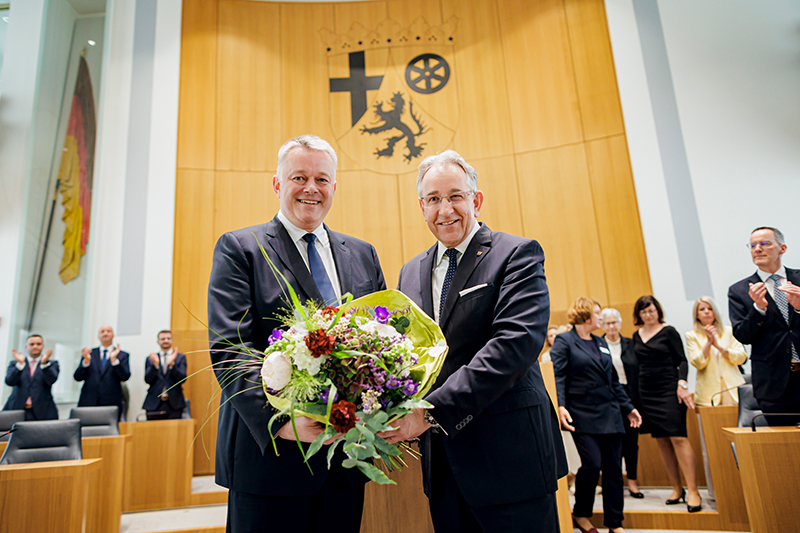 Gordon Schnieder und Matthias Lammert stehen nebeneinander im Plenarsaal und halten gemeinsam einen großen Blumenstrauß. Im Hintergrund applaudieren mehrere Personen.