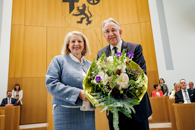 Marion Schneid und Matthias Lammert stehen nebeneinander im Plenarsaal und halten gemeinsam einen großen Blumenstrauß. Im Hintergrund sitzen mehrere Personen.
