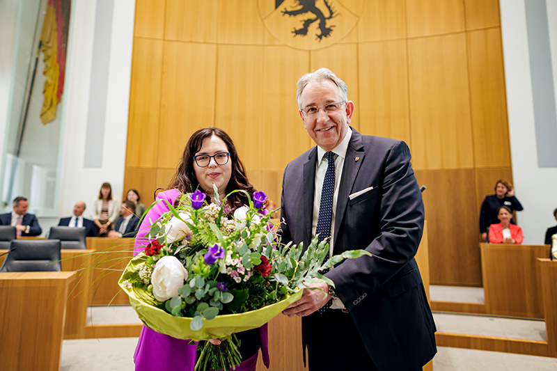 Jaqueline Rauschkolb und Matthias Lammert stehen nebeneinander im Plenarsaal und halten gemeinsam einen großen Blumenstrauß. Im Hintergrund sitzen mehrere Personen.