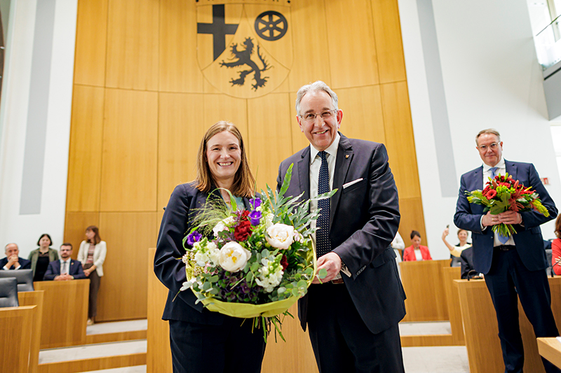 Katharina Binz und Matthias Lammert stehen nebeneinander im Plenarsaal und halten gemeinsam einen großen Blumenstrauß. Im Hintergrund sitzen mehrere Personen.