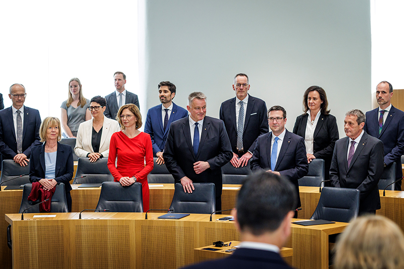 Die Staatsministerinnen und Staatsminister stehen gemeinsam mit Gordon Schnieder hinter ihren Plätzen im Plenarsaal.