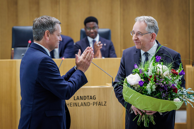 Zwei Männer in Anzügen stehen im Plenarsaal. Der Mann rechts hält einen großen Blumenstrauß und lächelt.