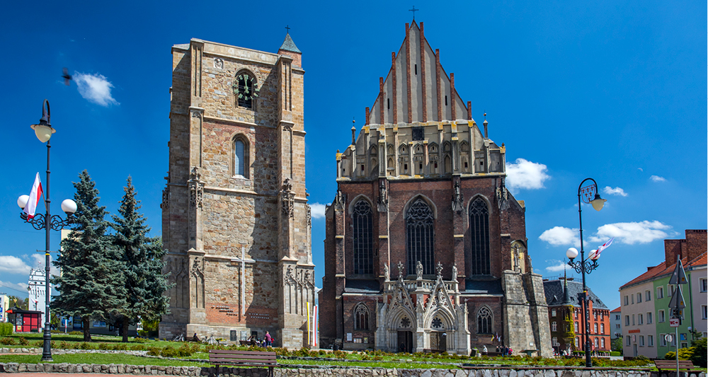 Zu sehen ist eine Kirche. Links im Bild befindet sich ein Glockenturm während das rechte Gebäude das gotische Hauptschiff der Basilika zeigt. Die gotische Fassade der Basilika ist durch hohe, spitz zulaufende Fenster, Fialen (Ziertürmchen) und eine imposante Eingangspartie mit reich verzierten Portalen und Figuren gekennzeichnet. Die Basilika ist umgeben von Bäumen und Laternen.