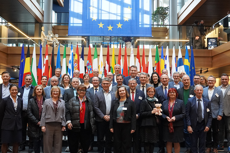 Auf dem Bild sieht man eine große Gruppe von Menschen in einem modernen Gebäude, die für ein Gruppenfoto posieren. Im Hintergrund hängen zahlreiche Nationalflaggen europäischer Länder sowie eine große Flagge der Europäischen Union.