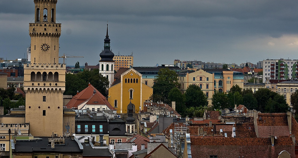 Auf dem Bild sieht man die Stadt Opole in Polen. Auffällig ist der Rathausturm im venezianischen Stil links im Bild, ein Wahrzeichen der Stadt. Im Hintergrund sind weitere Kirchen, Wohnhäuser und moderne Gebäude zu erkennen. Die Szenerie zeigt eine Mischung aus historischer und moderner Architektur.