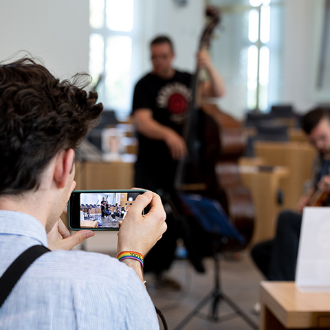 Zu sehen ist ein Besucher, der mit seinem Smartphone Musiker im Hintergrund aufnimmt.