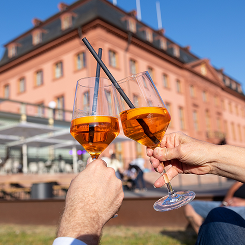 Zu sehen sind zwei Hände mit Aperol gefüllten Gläsern, die sich zu prosten. Im Hintergrund das Deutschhaus mit Restaurant. 