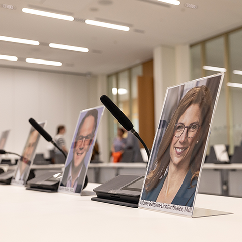 Zu sehen sind nebeneinder stehende Tische, auf denen Fotos von einigen Abgeordenten des Landtags stehen sowie dazwischenstehende Tischmikrophone.