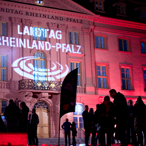 Zu sehen ist die Front des Deutschhauses bei Nacht. Die Hausfront wird mit buntem Licht angestrahlt, oberhalb des Eingangs wird das Logo auf die Hauswand projeziert. Im Vordergund sieht man einige Menschen stehen.