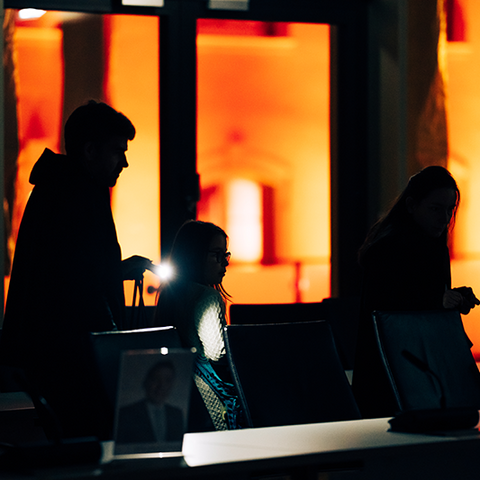 Zu sehen sind die Umrisse von drei Menschen, die durch einen Raum gehen. Im Hintergrund sieht man etwas Licht.