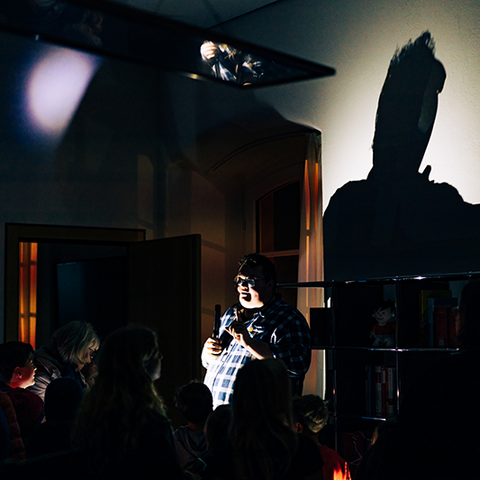 Zu sehen ist ein dunkler Raum. Mittig im Bild steht ein kräftiger Mann, der mit einer Taschenlampe angeleuchtet wird. Sein Schatten wird in Übergröße an die Wand geworfen. Man sieht die Umrisse einiger Kinder.