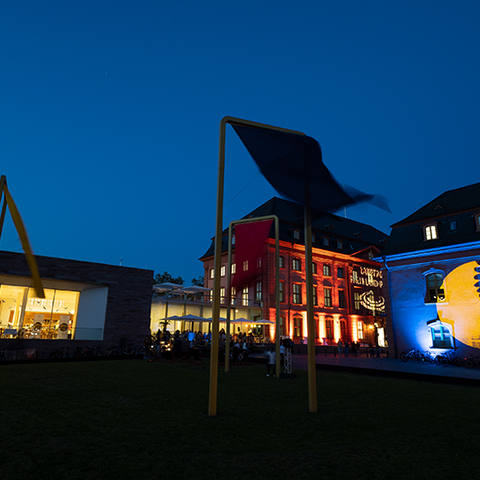 Zu sehen sind mehrere Gebäude bei Nacht, die angeleuchtet werden. Auf der rechten Seite sieht man das blau-gelbe Logo der Museumsnacht.